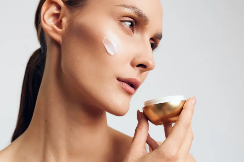 Young woman applies moisturizing cream on face holds jar with cosmetic product close to skin looks focused isolated on white background skincare beauty concept.