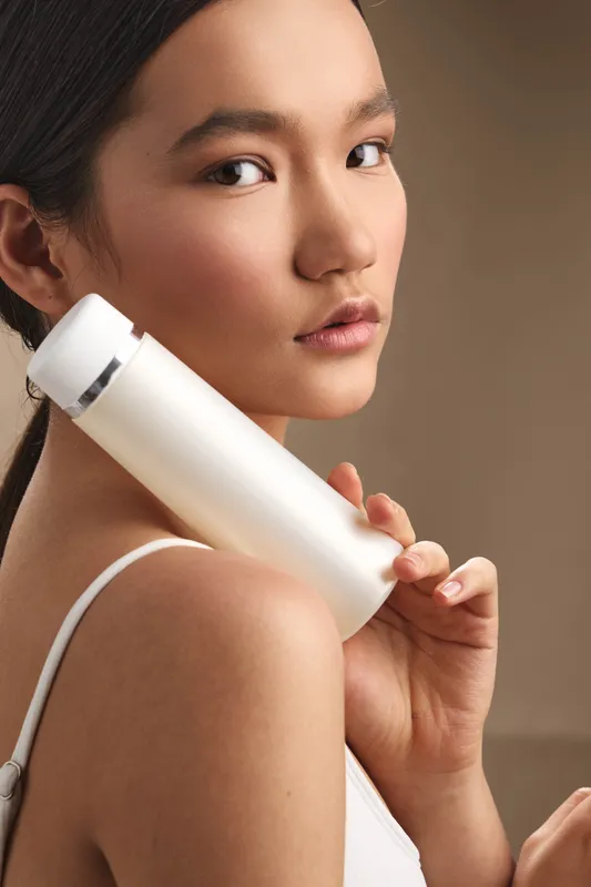 Asian woman holds a white skincare bottle near her shoulder, looking over her shoulder with a calm smile, showcasing a modern beauty routine in a softly lit, premium setting.