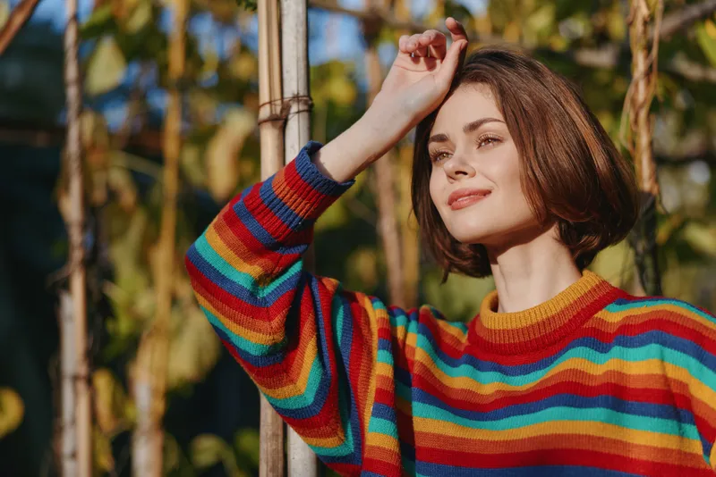 Woman in a colorful striped sweater with a short bob haircut, young adult smiling and leaning on a garden trellis outdoors in warm sunlight, lifestyle portrait for cozy fashion and travel.