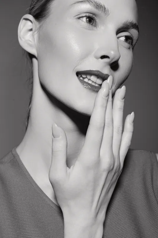Close-up of a young Caucasian woman with light skin touching her face, smiling gently. Concept of natural beauty, skincare, and self-care in studio lighting.