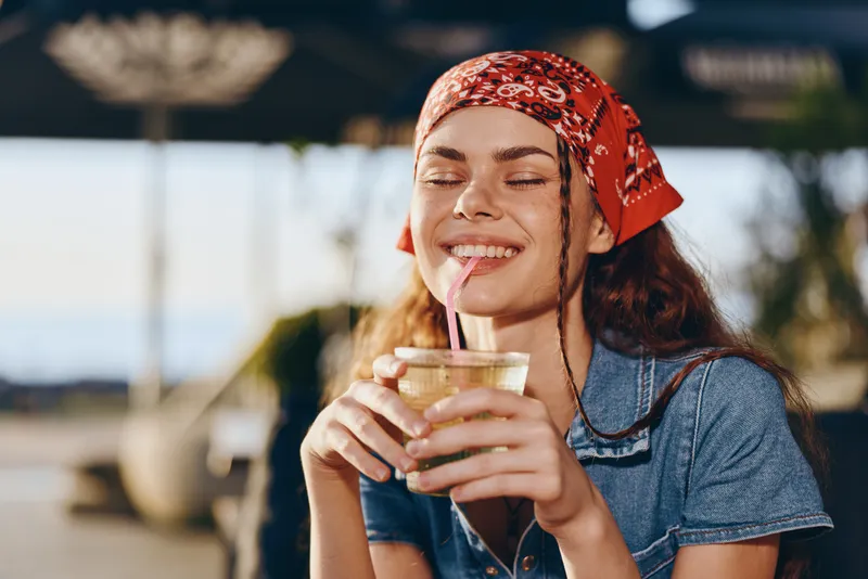 smiling woman enjoying refreshing drink with straw outdoors in summer wearing casual denim and red headscarf with relaxed expression and closed eyes