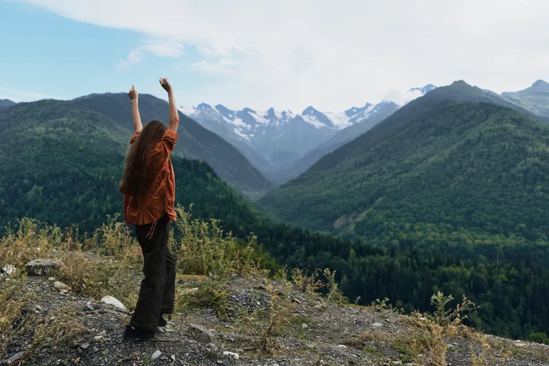 Joyful hiker stands on a rocky overlook with arms raised in triumph, savoring the expansive alpine landscape and distant snowy peaks under clear blue sky
