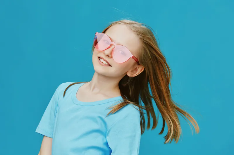 Happy young girl wearing pink sunglasses and a blue t shirt against a bright blue background, showcasing joyfulness and summer vibes