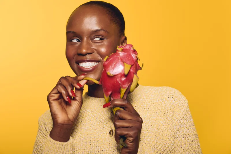 Smiling dark skinned African woman holding a ripe dragon fruit on a yellow background. Concept for healthy food, tropical fruit, nutrition, and positive lifestyle with natural beauty.