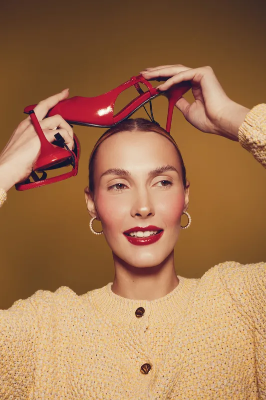 Young woman with red lipstick and hoop earrings holds red high heel shoes above her head. She wears a beige knitted sweater on a warm brown background. Concept of fashion and style.