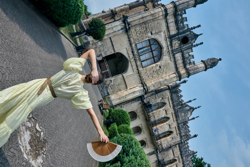 Dress clad woman strolls near a grand castle in a sunny garden holding a decorative fan, capturing historic architecture and outdoor travel vibes with graceful elegance