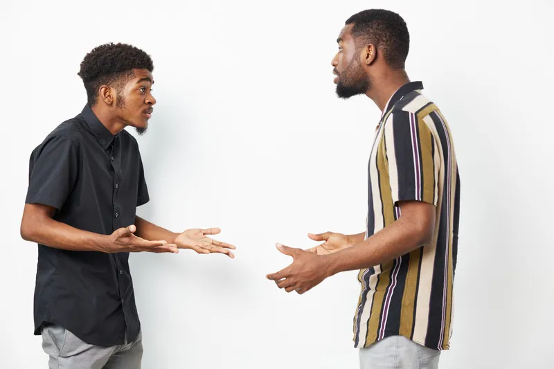 Two African American men engaging in a heated discussion, expressing strong emotions. The background is simple and neutral, allowing focus on their interaction.