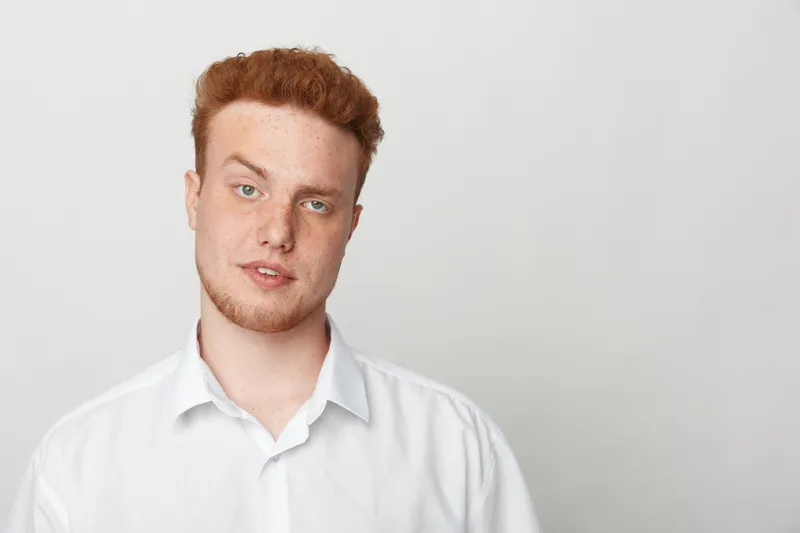 Young man with red hair and beard wearing white shirt looks at camera with neutral expression on light grey background. Concept of casual style, portrait, and natural appearance.