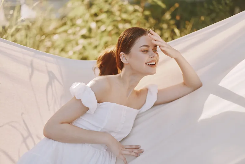 Beautiful young woman in a white dress smiling thoughtfully while relaxing on a hammock in a bright outdoor setting, evoking a sense of calm and joy