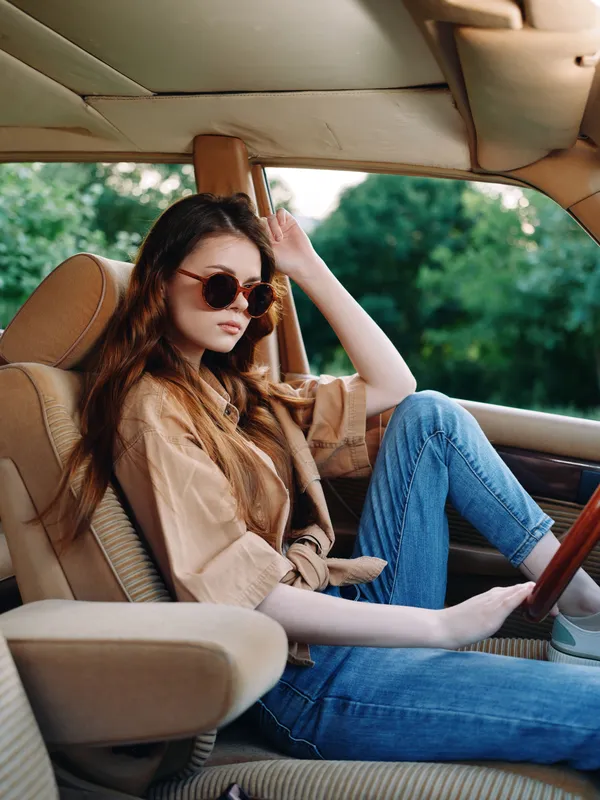 Stylish young woman in sunglasses relaxes in a vintage car, showcasing a trendy casual outfit, evoking a sense of freedom and adventure with a natural outdoor background.
