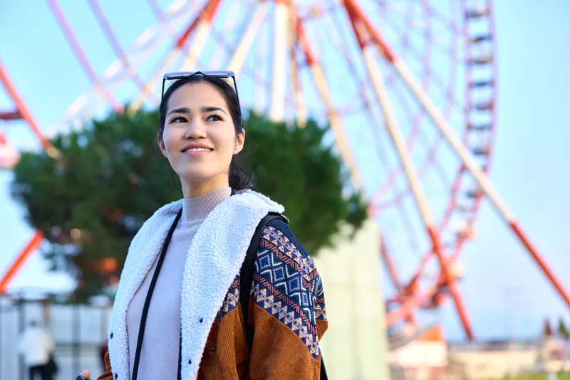 Woman near a large ferris wheel at an amusement park, outdoor scene, smiling in a cardigan and scarf, casual chic, sunny day, capturing a joyful leisure moment