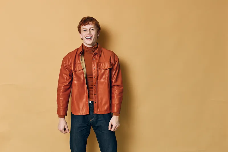 Young man standing and laughing in casual outfit with brown leather jacket and jeans against beige background. Happy adult male showing joy and relaxed posture.
