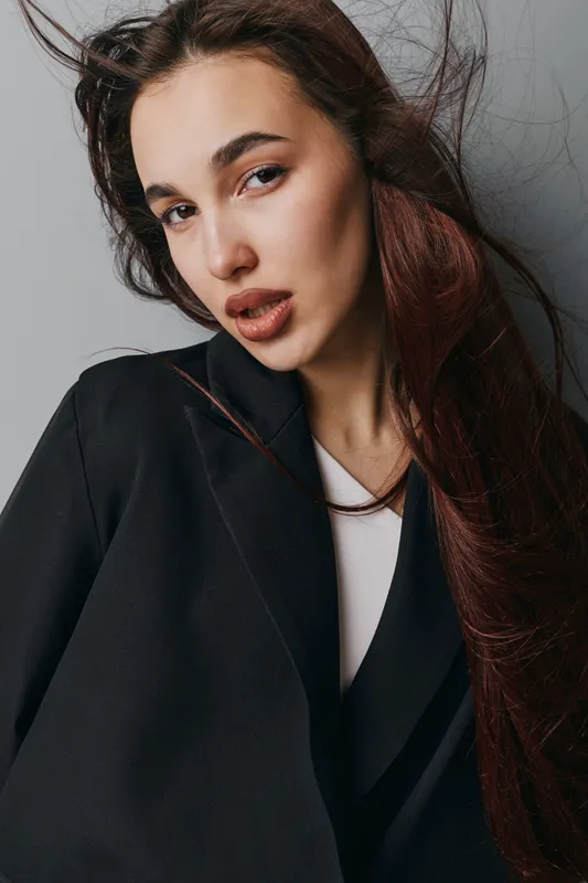 Portrait of young woman with long hair wearing black coat and white shirt against gray background. Fashion model posing with natural makeup and serious expression in studio light.
