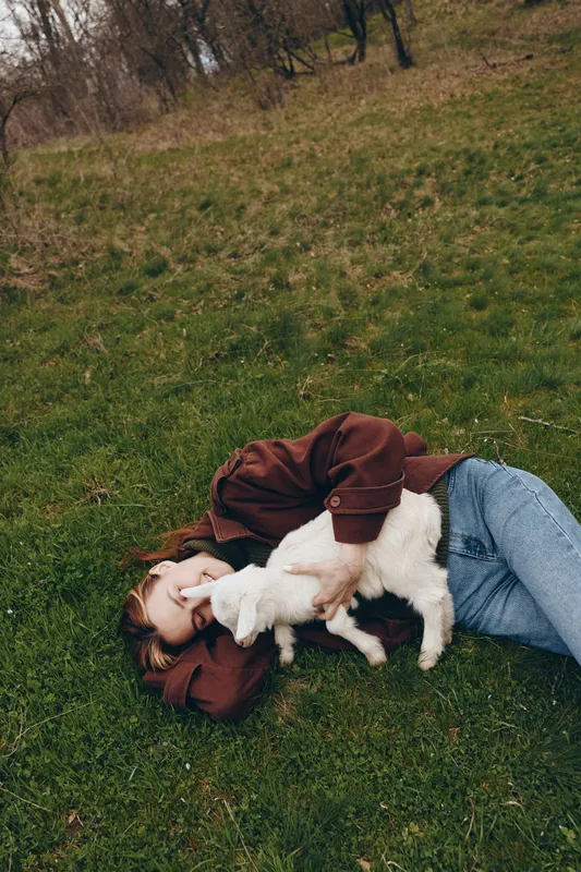 Young woman lies on grass in casual jacket and jeans, cuddling a gentle goat kid, sharing a tender moment in nature, outdoor lifestyle scene with peaceful rural ambiance