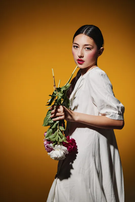 Asian woman with sleek hairstyle poses holding flowers wearing white dress against orange background. Creative beauty and fashion shot highlighting natural makeup and soft light.