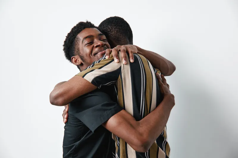 Two young black men hugging each other with smiles and closed eyes for friendship and support. African ethnicity, dark skin, casual clothing, positive emotion and connection.