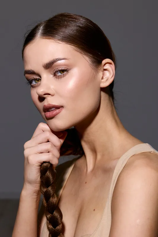 Beauty woman with long braided hair and flawless skin posing against a dark background, showcasing natural makeup and soft lighting for a fashion portrait.