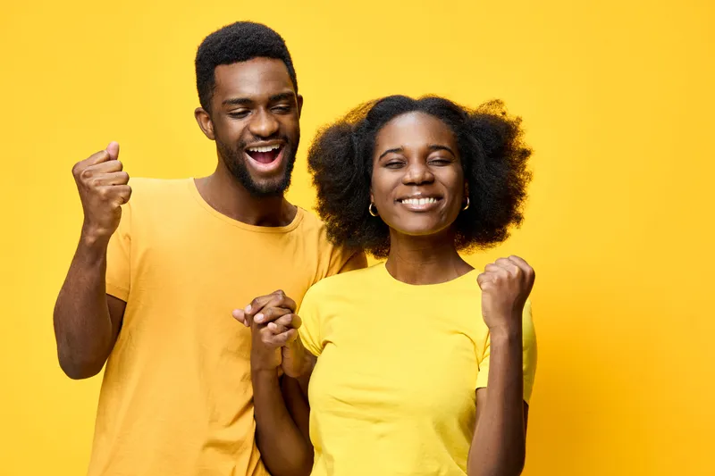 Happy young black couple celebrating together in matching yellow t shirts on a bright yellow background, showing joy and excitement