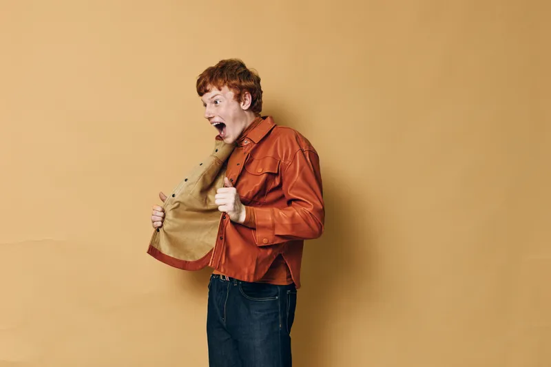 Man with surprised expression opening jacket in excitement, wearing orange shirt and dark jeans on beige background, showing emotion and casual style.