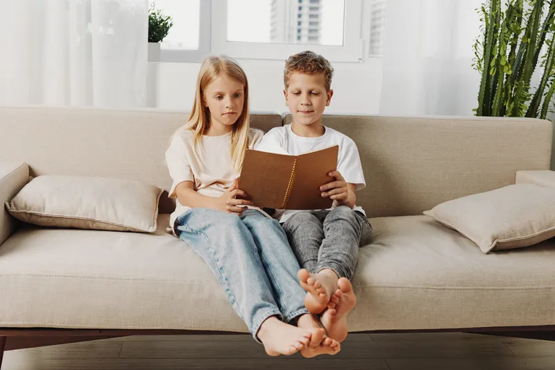 Two barefoot children sitting closely on a beige sofa reading a book together in a bright living room with modern decor and plants. Concept of childhood learning and family bonding.