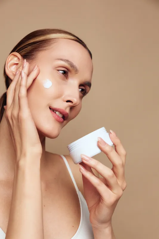 Skincare beauty woman face cream moisturizing cosmetics selfcare routine spa concept, young woman applies moisturizing cream with cotton pad, gentle expression, beige background