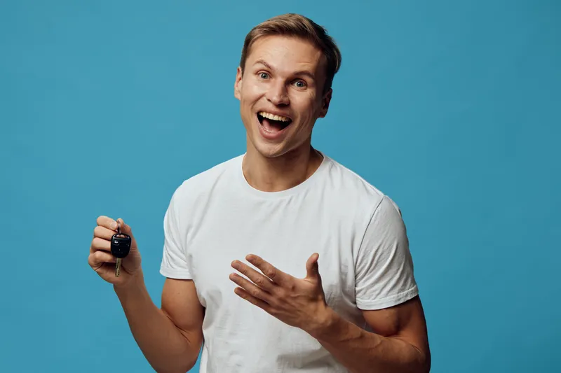 Joyful smiling man holding car key in hand looking at camera, wearing casual white t-shirt, emotional expression, studio portrait with solid light blue background, lifestyle concept, happiness.