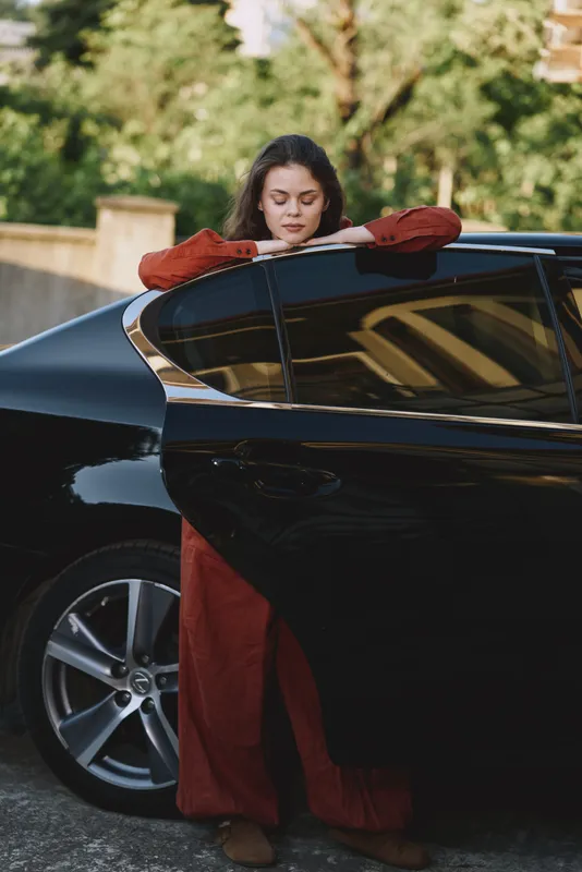 Young woman in a red coat leaning on a sleek car door in an outdoor setting. Lifestyle scene focused on fashion, confidence, and everyday travel moments