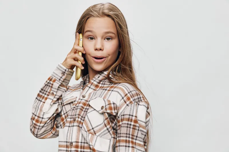 Girl with long hair speaking on smartphone, wearing checkered shirt with buttons, standing against light background with casual expression and hand near face.