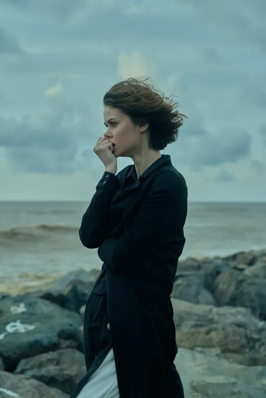 Coastal wind, thoughtful mood, outdoor scene with a coat at the shoreline, gazing toward the ocean as waves crash against rocks and mist drifts over the horizon.
