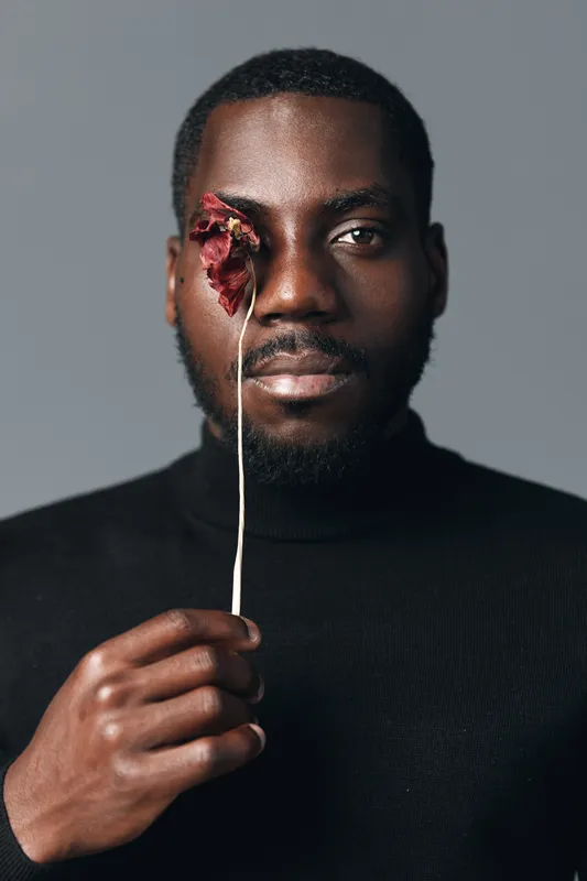 Creative conceptual portrait of African man holding dried flower in front of eye with serious expression, minimalistic visual style and dark clothing on gray background.