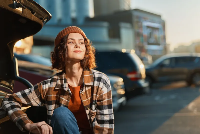 Woman portrait by car trunk in urban parking during golden hour glow sunlight, wearing beanie and plaid jacket, candid lifestyle authenticity, mindful living and emotional storytelling mood.