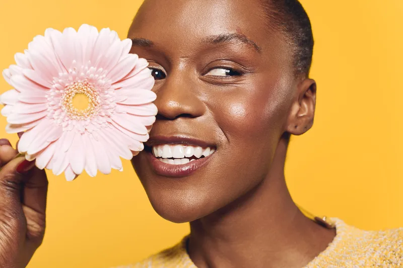 Close-up of happy African woman with dark skin holding pink flower near face smiling on yellow background. Concept of natural beauty, skincare, wellness, and positive mood.