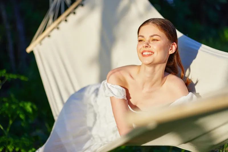 Relaxed young woman enjoying sunny day in hammock, wearing a white dress, embodying happiness and tranquility in a lush green environment
