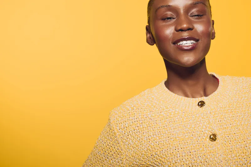 Smiling African American woman with dark skin wearing a yellow knitted sweater poses against a bright yellow background. Concept of happiness, fashion, and confident beauty.