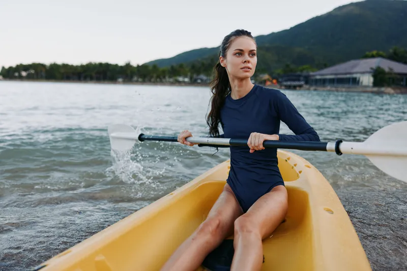 A focused female kayaker sits in a yellow kayak, gripping a paddle as water splashes, ready to glide across calm lake under clear sky.
