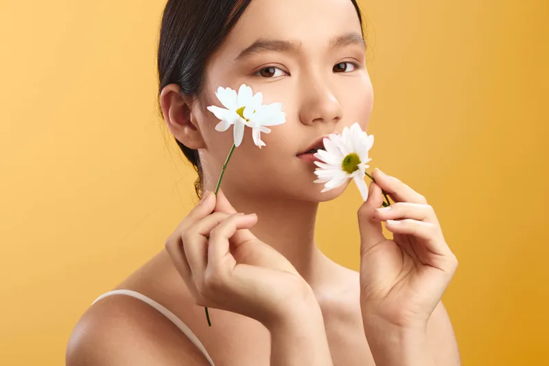 asian woman holds white daisies near her face against a warm yellow background. soft lighting enhances a fresh beauty portrait with natural makeup and a serene, artistic vibe.