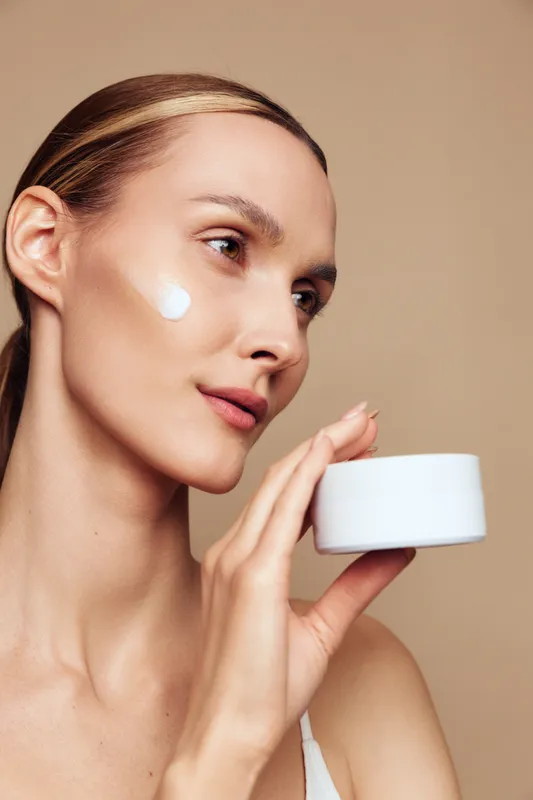 Young woman with fair skin applies facial cream to her cheek in a neutral studio, calm focused expression captures a intimate skincare moment
