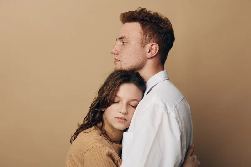 Young couple embraces tenderly against beige background. Woman with closed eyes rests head on man’s chest. Concept of love, comfort, support, intimacy, and emotional connection.