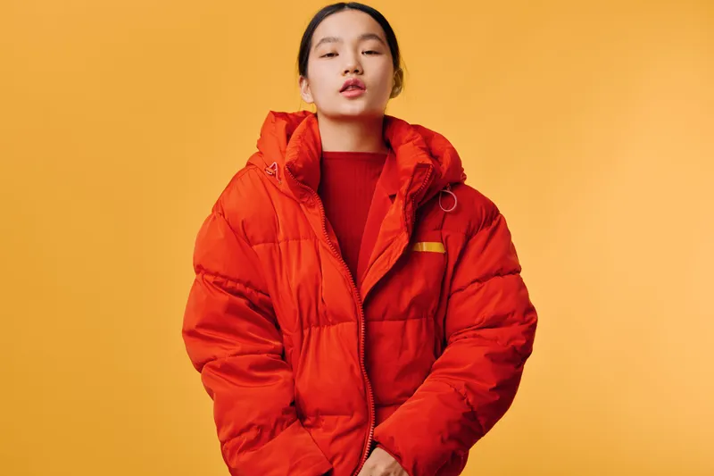 Young woman wearing orange puffer jacket with high collar standing against yellow background looking confident and stylish casual fashion portrait with winter outerwear and minimal studio lighting.