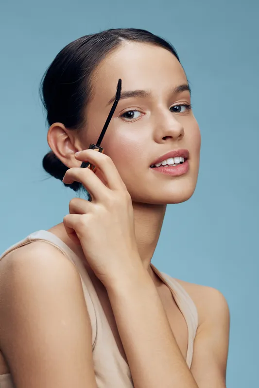 Young woman applying brow gel with a confident smile, showcasing beauty and confidence against a soft blue background Makeup, beauty, confidence, elegance, model