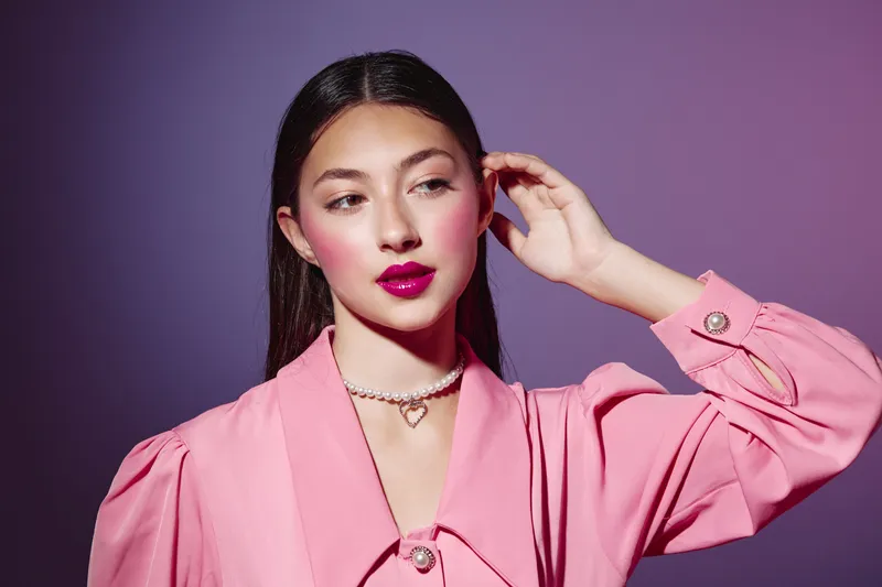 Young woman in a pink trendy outfit with bright makeup poses thoughtfully against a purple background. Concept for fashion, beauty, and style innovation.