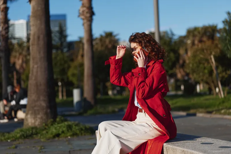 A woman in a red coat sits on a park bench, talking on a smartphone, shielding her eyes from sunlight. An outdoor urban park scene highlighting casual style and everyday communication.