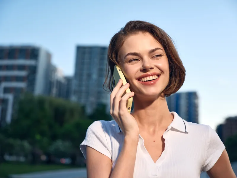 A cheerful woman talks on a phone outdoors in a modern city setting. She smiles proudly, creating a vibrant and professional atmosphere.