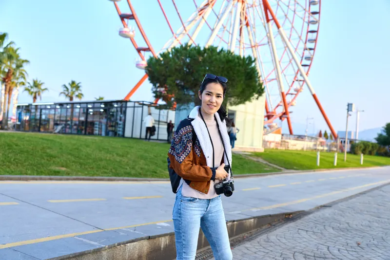 Woman with camera stands near a large ferris wheel in a sunny park, casual denim and scarf, smiling as she captures outdoor life, travel vibes, and cheerful street photography moments