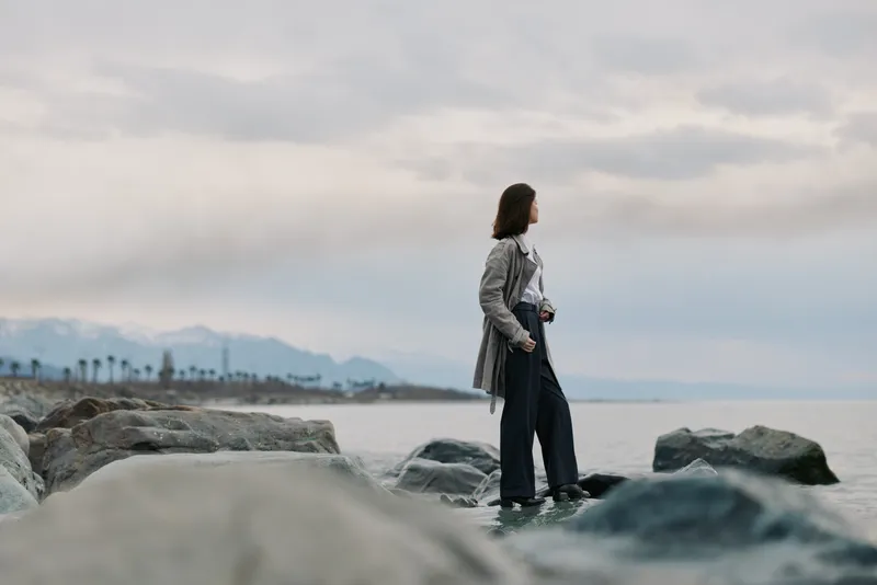 Young woman stands on rocky shore looking at calm sea under cloudy sky. She wears casual clothes and holds a camera, creating a contemplative and peaceful mood by the water.