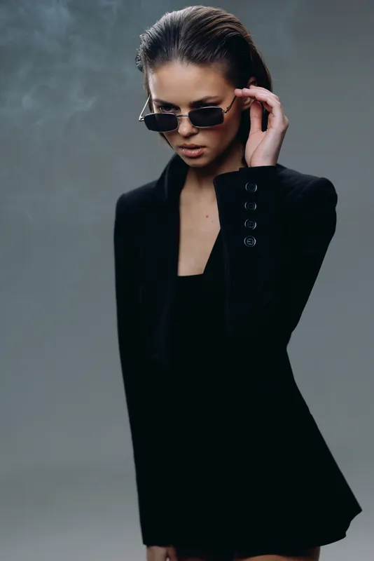 Stylish woman in black outfit wearing sunglasses with serious expression and smoky background