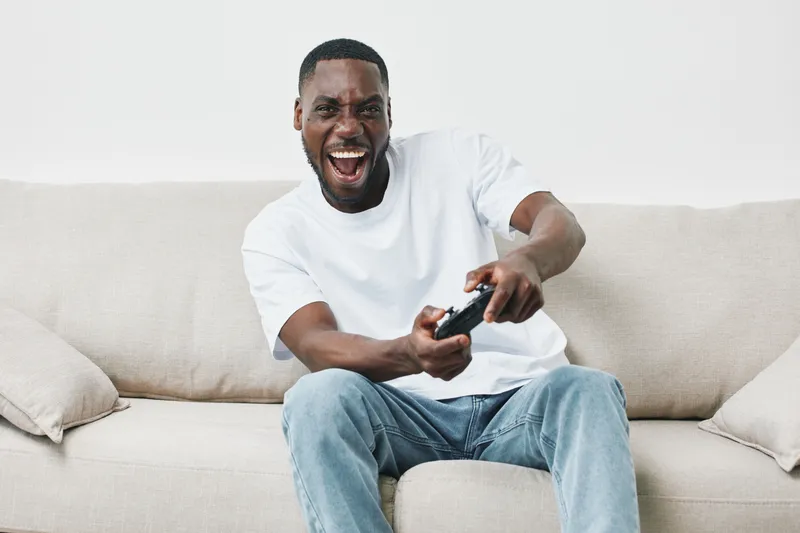 Happy young African American man playing video games on a couch, showcasing excitement and joy with a game controller in a cozy living room setting