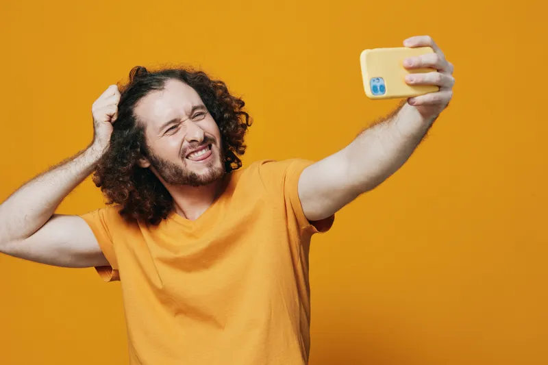 Young man with curly hair taking a selfie on a vibrant yellow background, expressing joy and confidence in casual clothing