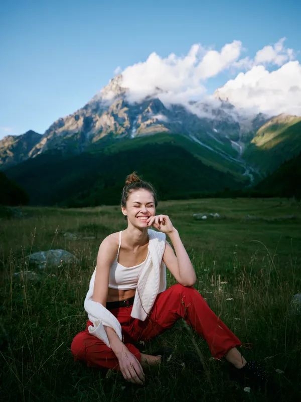 Outdoor landscape scene shows a smiling woman resting in a meadow with mountains in the distance, casual attire, relaxed pose, and serene scenery for travel or lifestyle stock