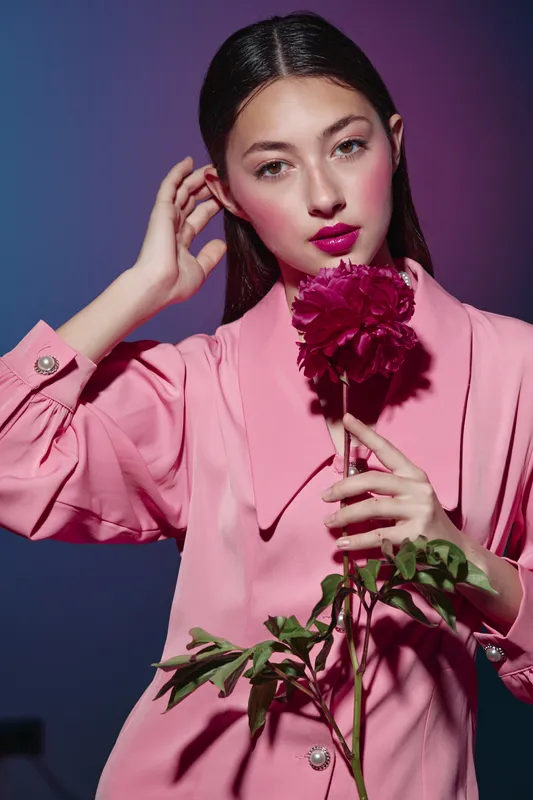Young woman in pink coat holding a dark pink flower close to her face, posing against purple gradient backdrop. Beauty and fashion creative concept with vibrant color tones.